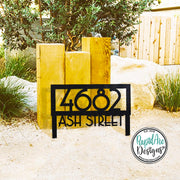 May include: Black metal address sign displaying the numbers "4682" and the words "ASH STREET". The sign is in front of three wooden posts and a large rock. The sign is in a garden setting.
