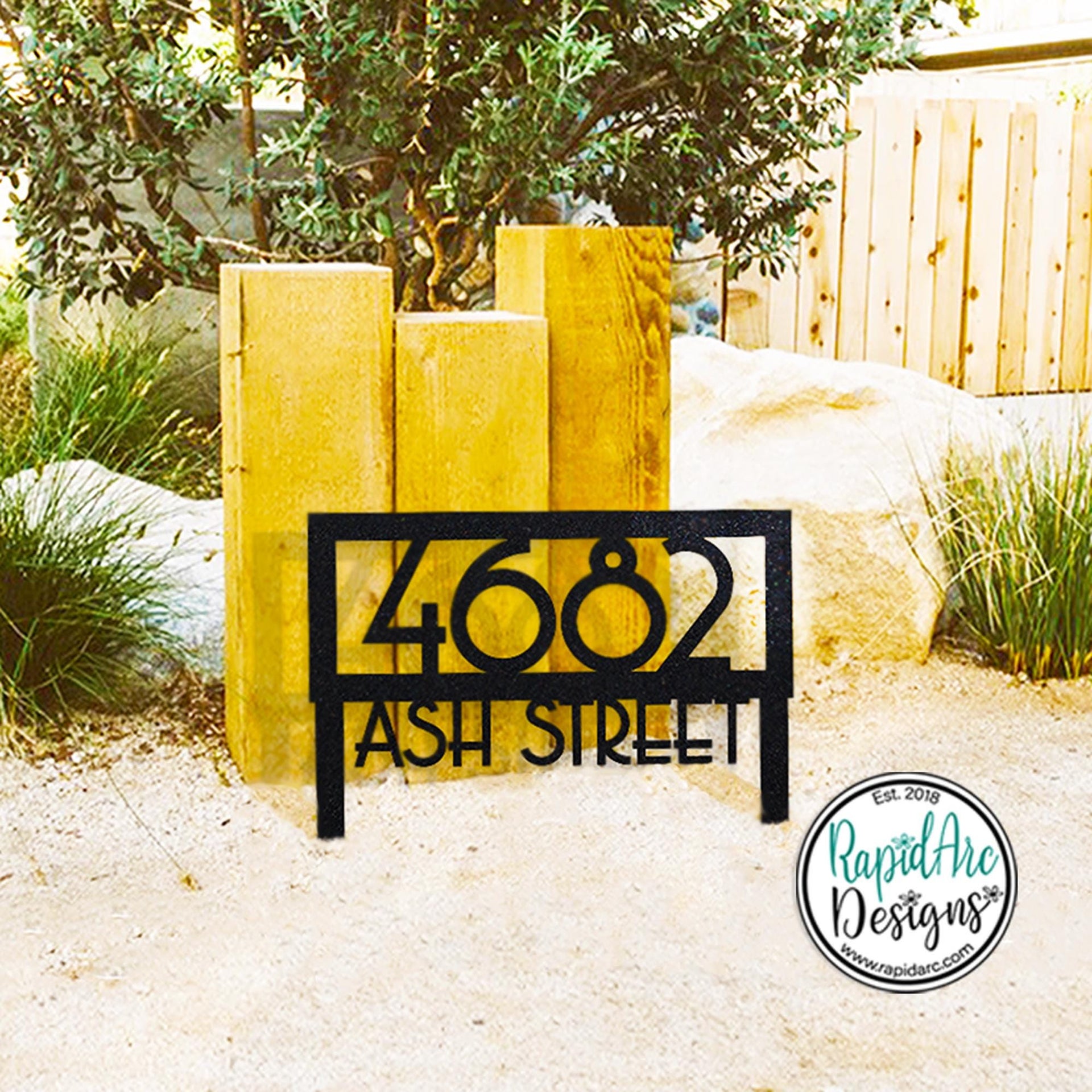 May include: Black metal address sign displaying the numbers "4682" and the words "ASH STREET". The sign is in front of three wooden posts and a large rock. The sign is in a garden setting.